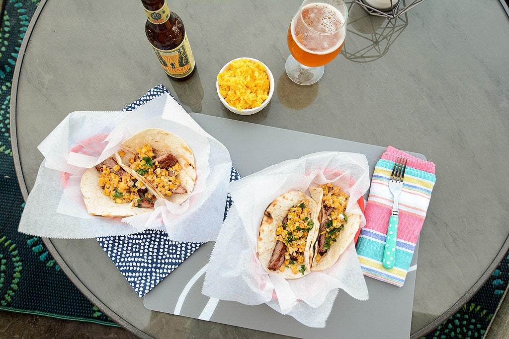 Overhead photo of Grilled Carne Asada Tacos and beers and corn salad on a table