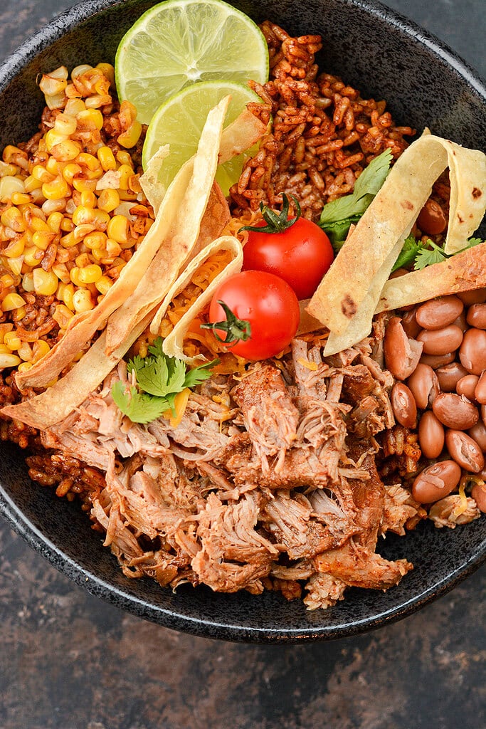 Pulled Pork over rice with corn and beans in black bowl topped with limes and tomatoes.