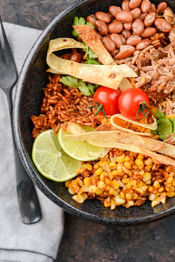 Overhead view of pulled Pork over rice with corn and beans in black bowl topped with limes and tomatoes.