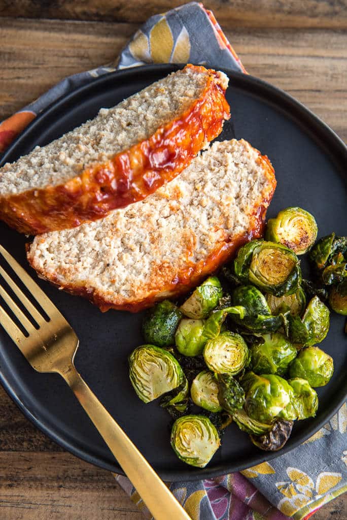 Sliced turkey meatloaf on a black plate with Brussels sprouts and a gold fork.