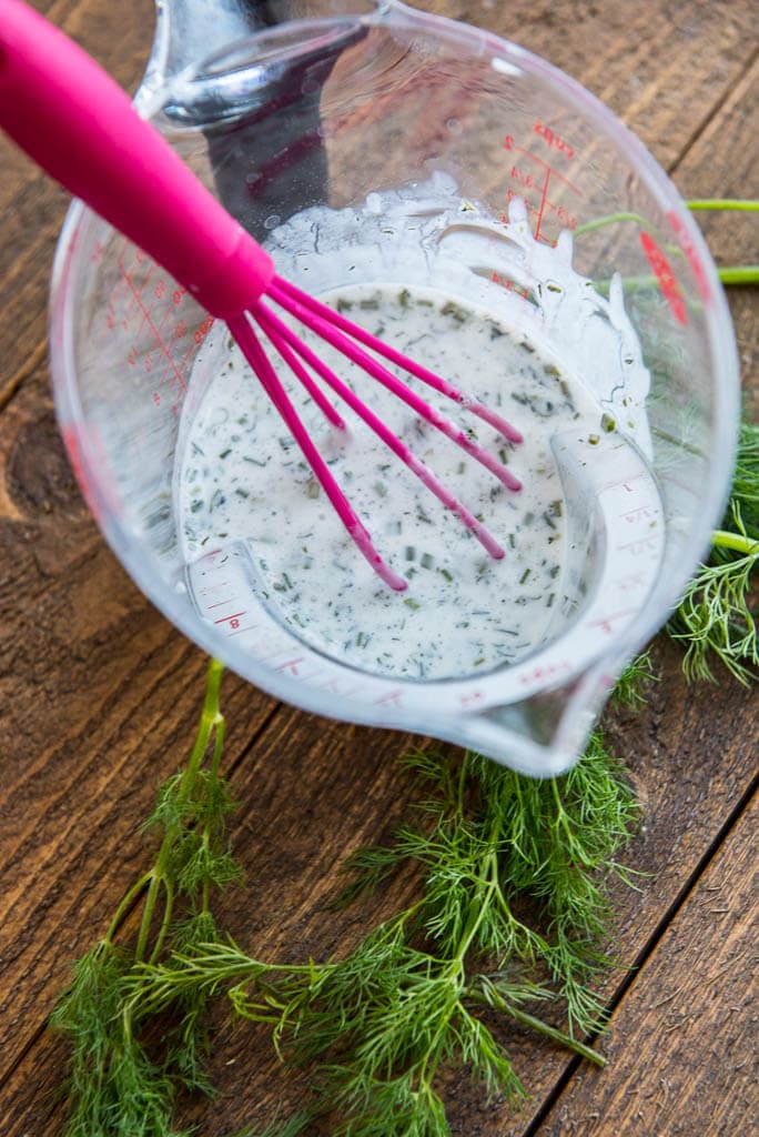 Homemade buttermilk dressing with a pink whisk and fresh dill for Wedge Salad with Herbed Buttermilk Dressing.