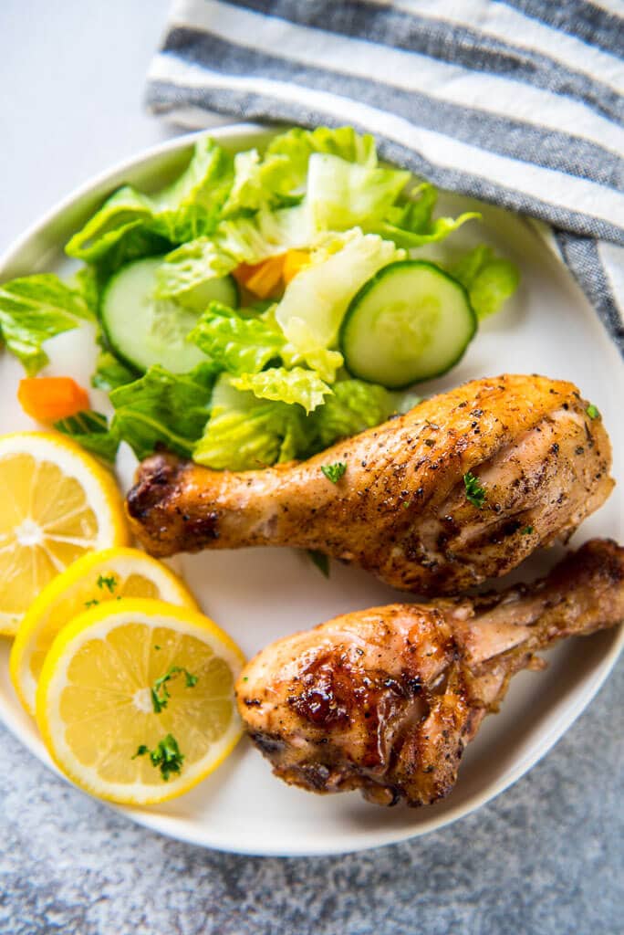 two Lemon Herb Grilled Chicken Drumsticks on a white plate with sliced lemons and a green tossed salad