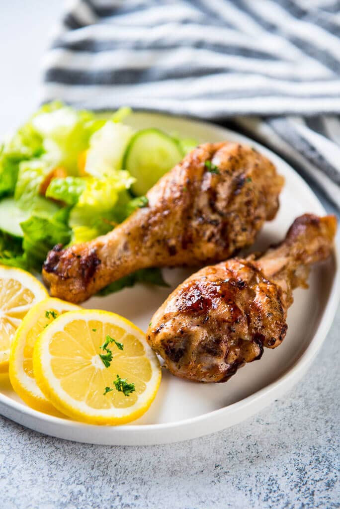 two Lemon Herb Grilled Chicken Drumsticks on a white plate with sliced lemons and a green tossed salad