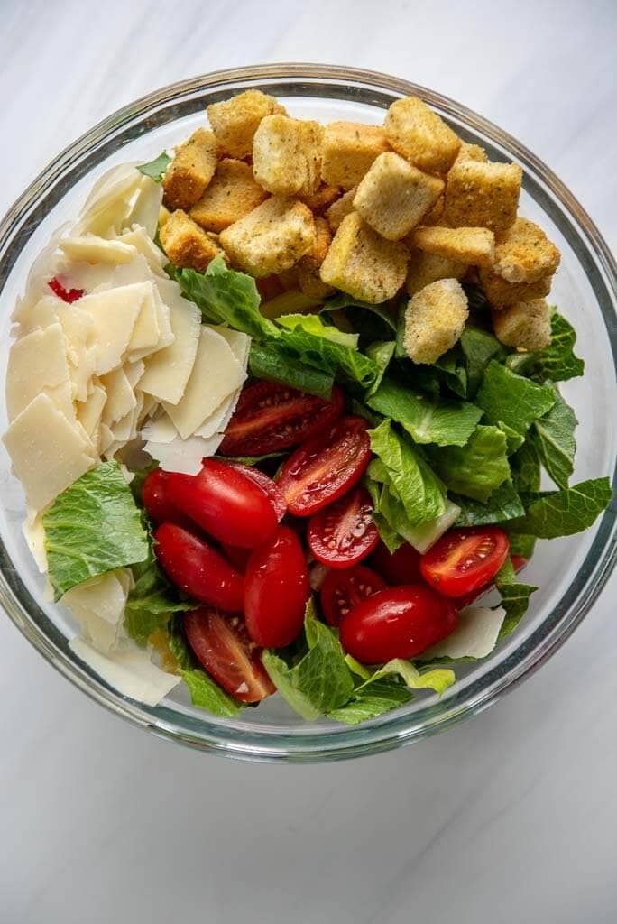 tomatoes, lettuce, croutons and cheese atop pasta in a bowl to mix for Caesar pasta salad 