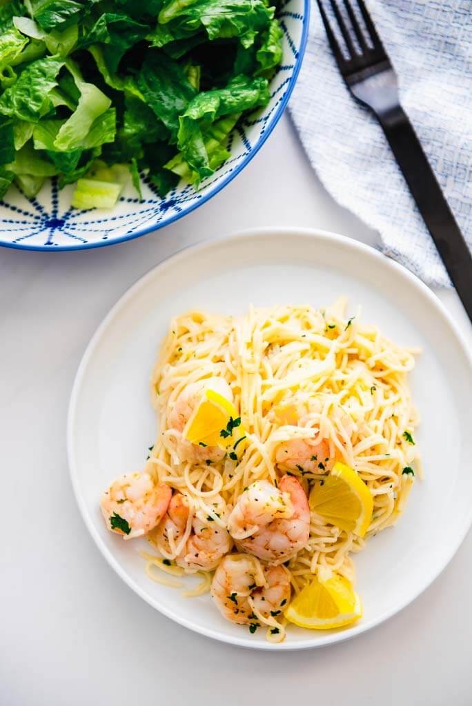 Lemon Garlic Shrimp Pasta on white plate with bowl of salad and a black fork on a white and blue napkin