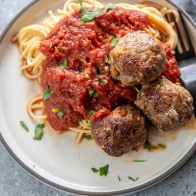 white plate with spaghetti noodles, marinara sauce, 3 meatballs and garnished with chopped parsley