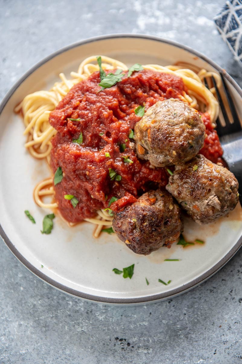 white plate with spaghetti noodles, marinara sauce, 3 meatballs and garnished with chopped parsley