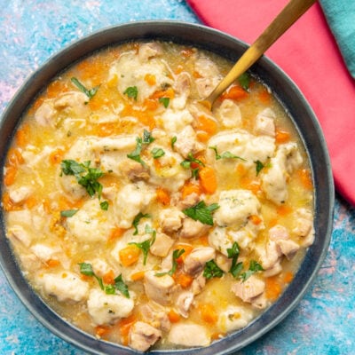 blue bowl filled with chicken and dumplings on a blue splattered backdrop with a pink napkin