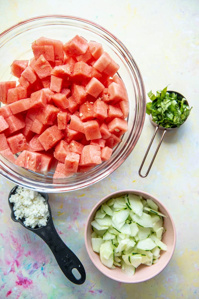 bowl of watermelon cubes, bowl of sliced cucumber, tablespoon of basil and tablespoon of cheese