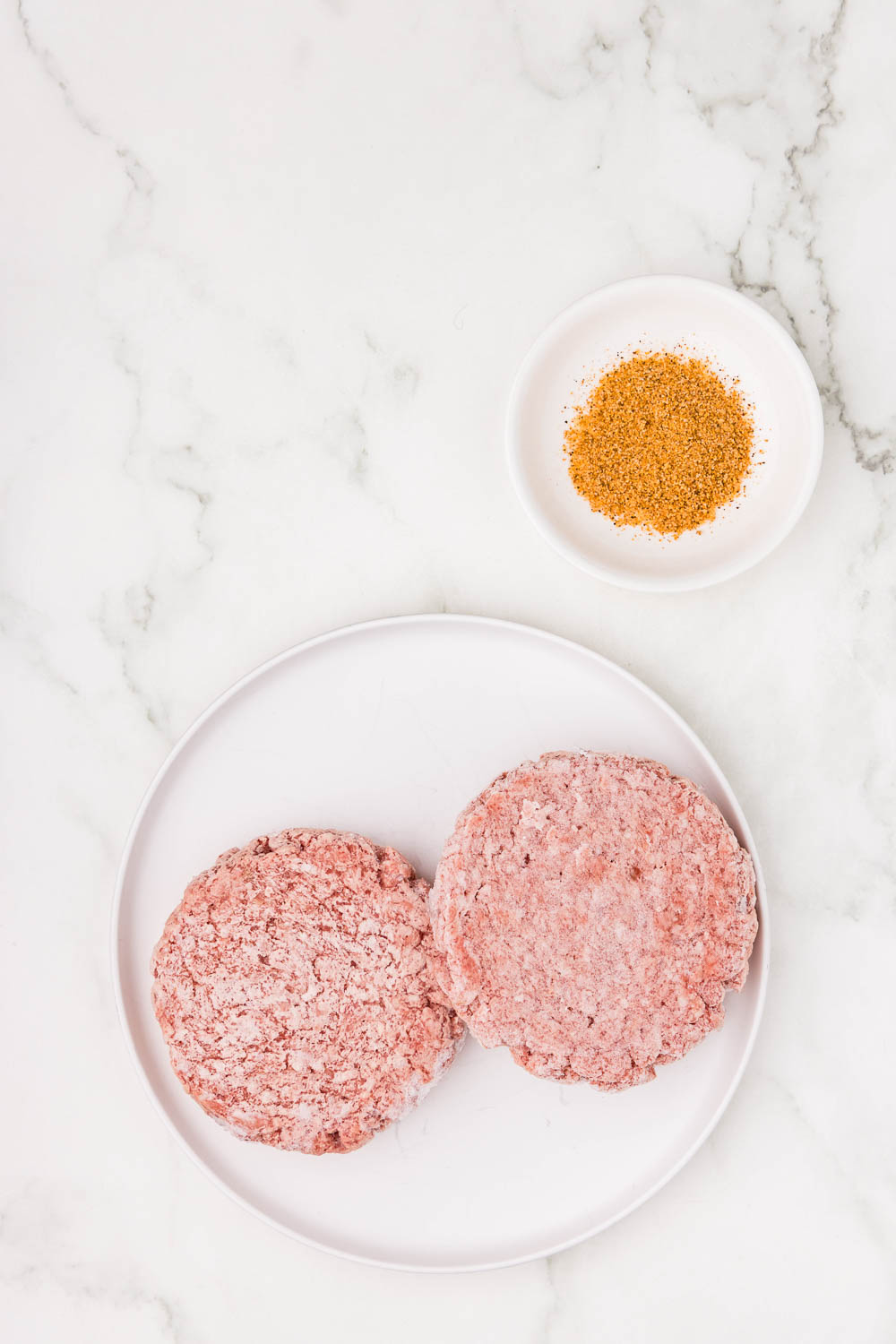 2 uncooked burger patties on dish next to small dish with seasoning