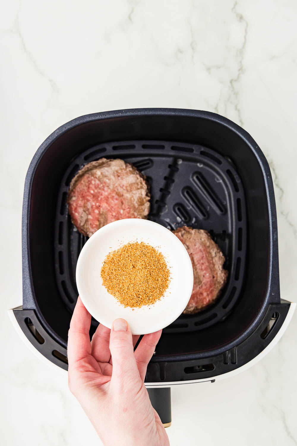 2 burger patties uncooked in air fryer and a dish of seasoning being held on top