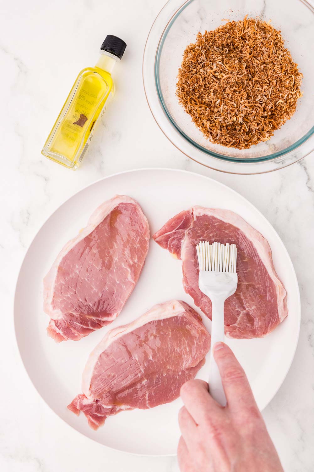 3 uncooked boneless pork chops being brushed with oil