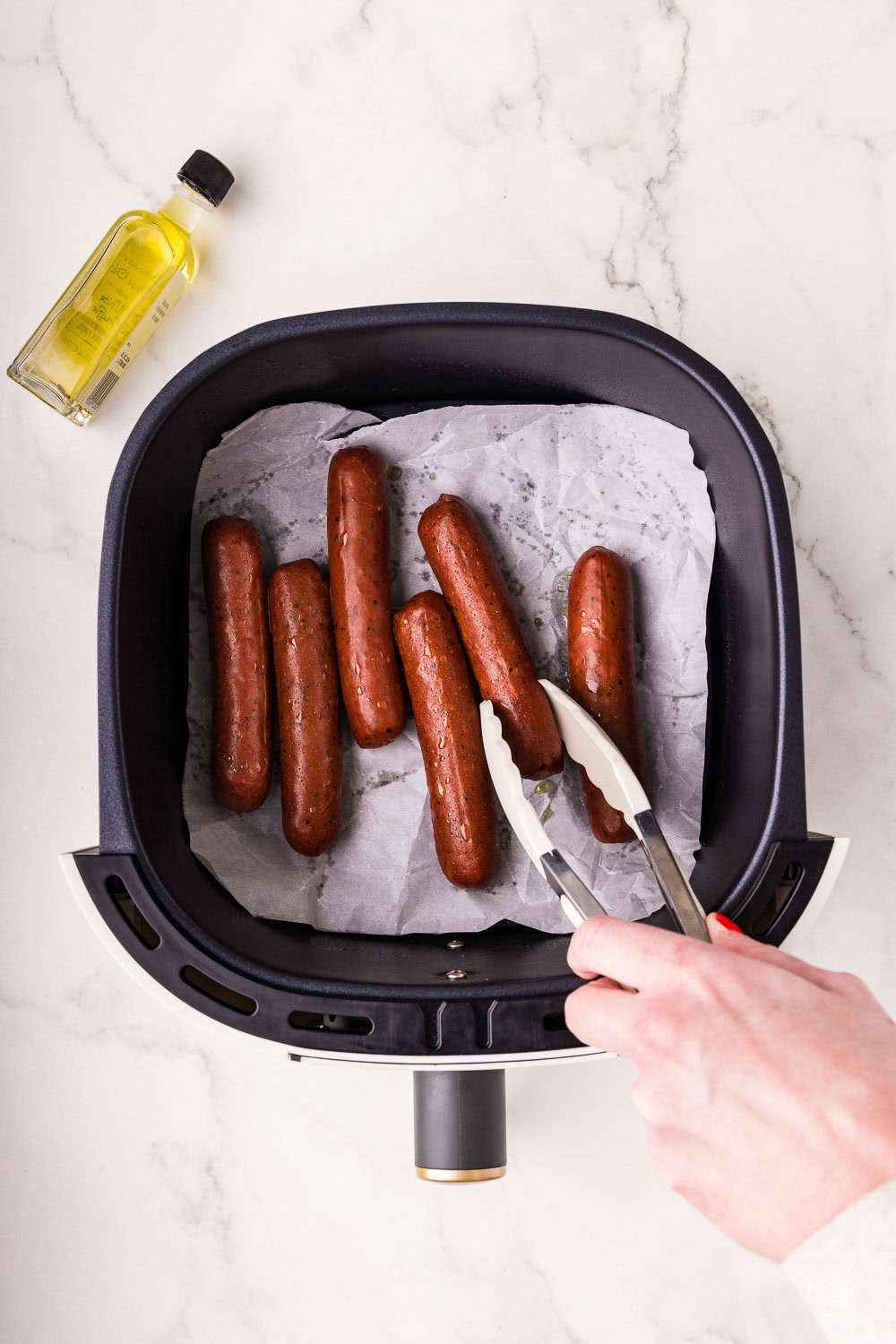 Chicken sausage on parchment paper in air fryer and tongs picking up a sausage to flip
