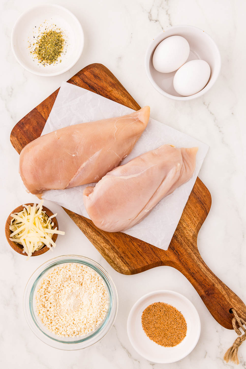 2 chicken cutlets on cutitng board next to panko crumbs, seasonings, cheese