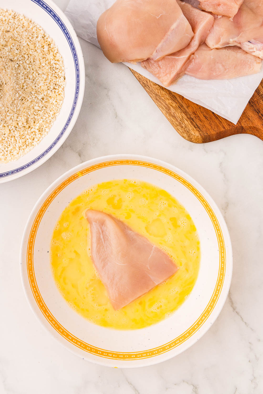 Chicken in dish with beaten egg, panko breadcrumbs, next to raw chicken cutlets