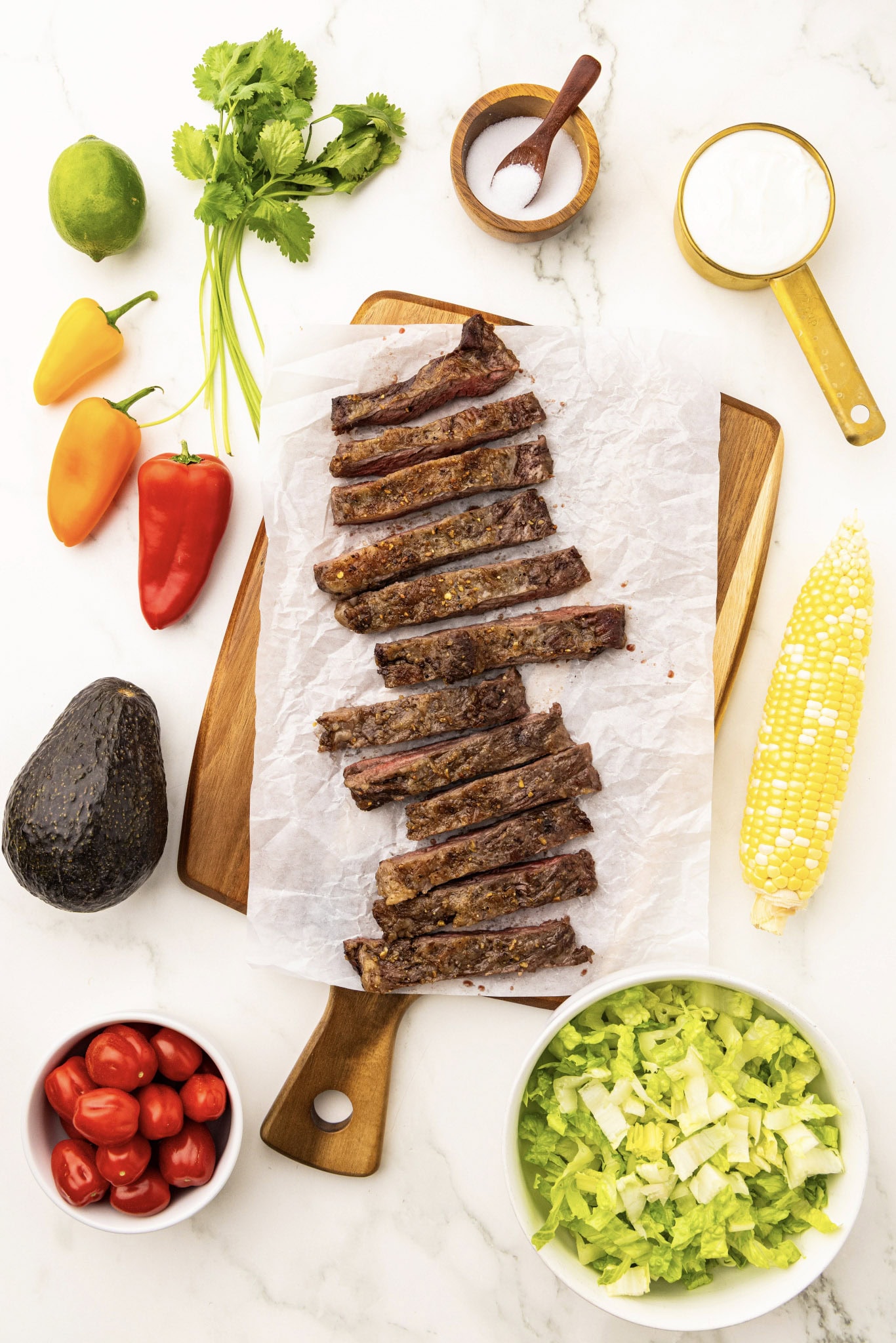 A cutting board with steak, corn, tomatoes, avocado and other ingredients