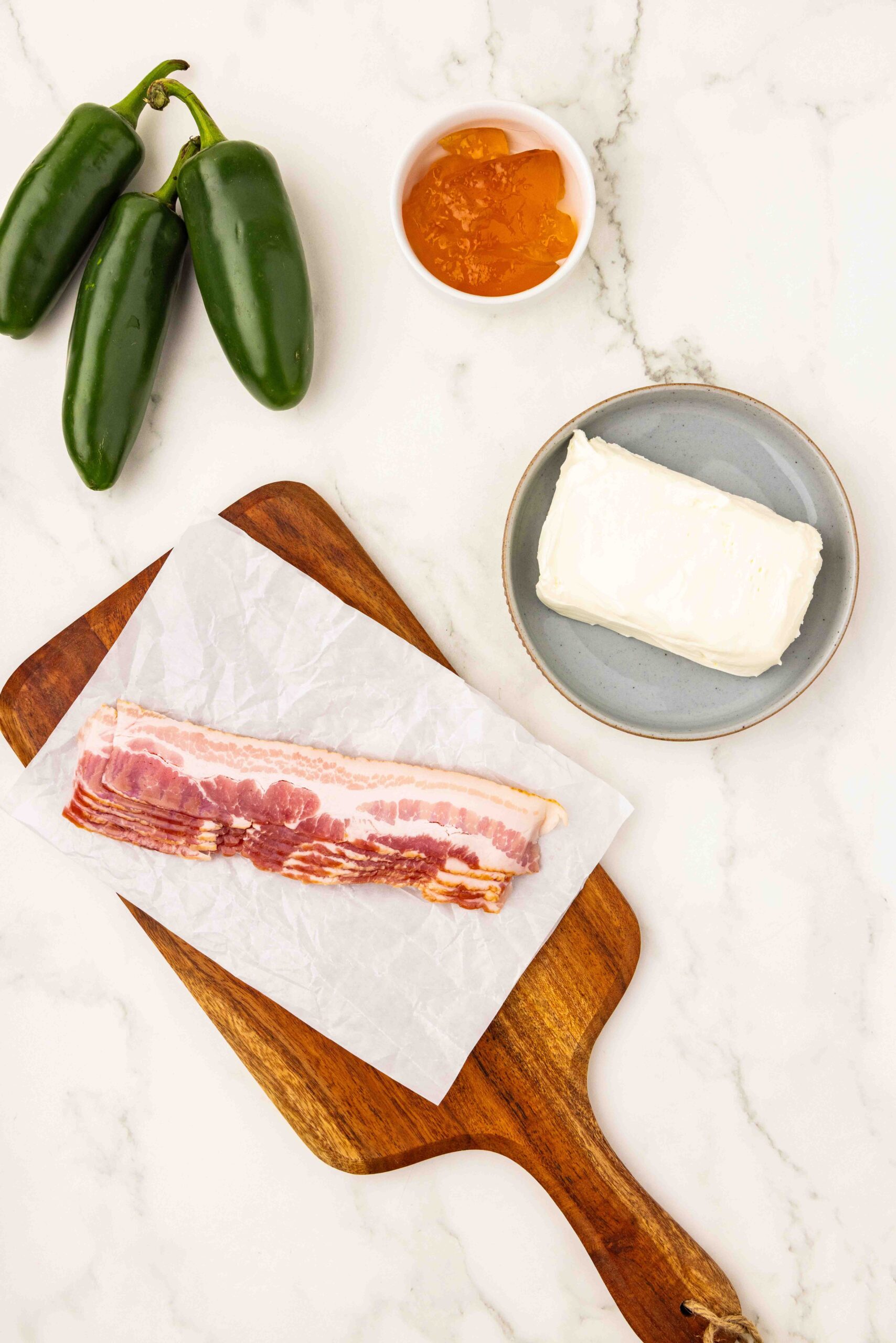 Bacon slices on top of cutting board next to cream cheese, jam, and jalapeños