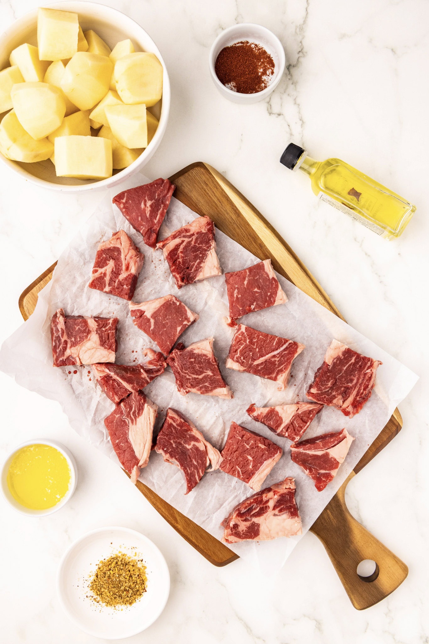 cut up steak on parchment and wooden cutting board, surrounded by bowls of cut up potato, butter, seasonings and olive oil