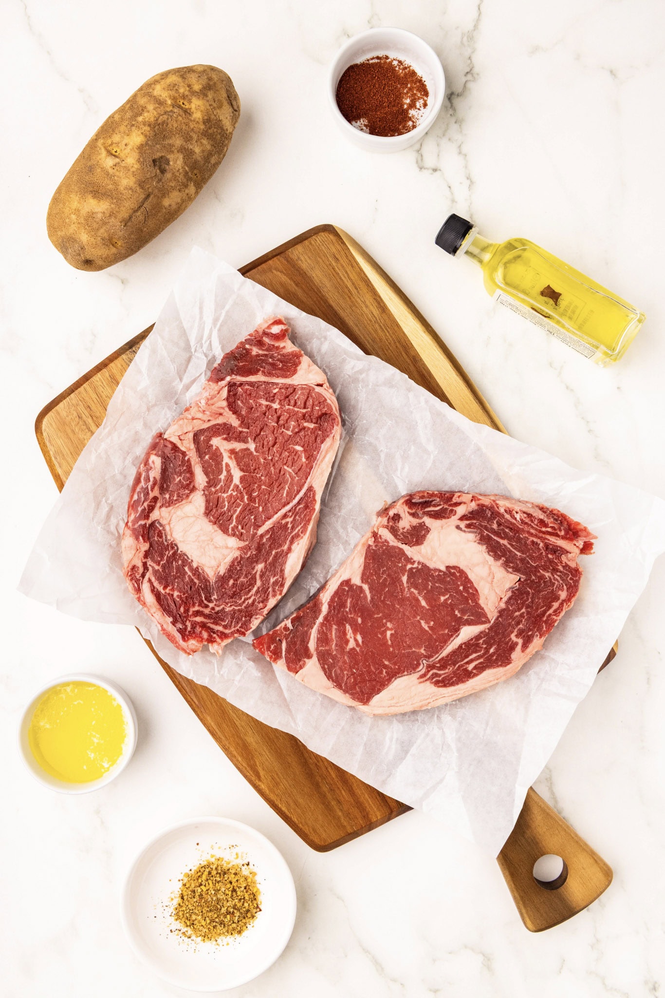 ingredients to include two ribeye steaks on parchment paper and wooden cutting board, surrounded by one russet potato, bottle of olive oil, and bowls of steak seasoning, melted butter and paprika
