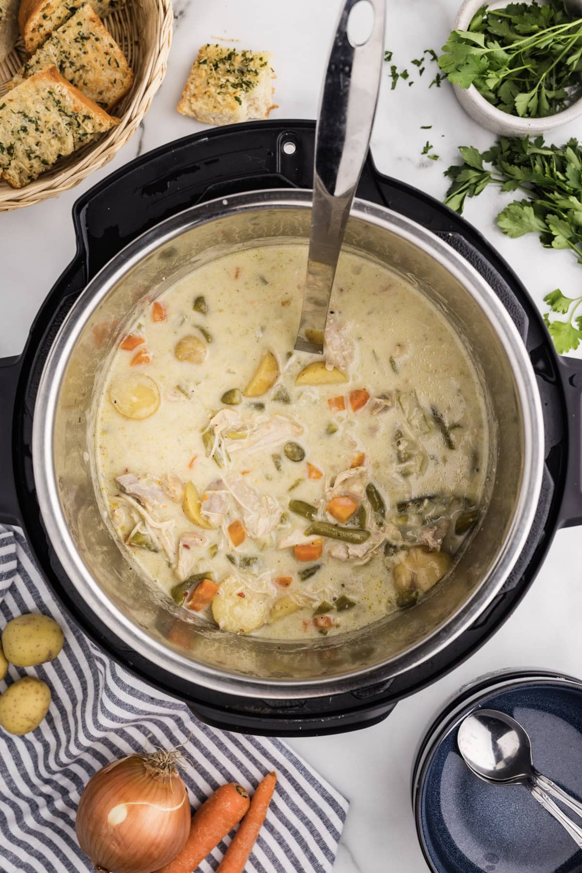 overhead shot of instant pot with soup ladle, with bowl of fresh parsley, basket of bread, soup bowls and spoons, and assorted veggies surrounding instant pot