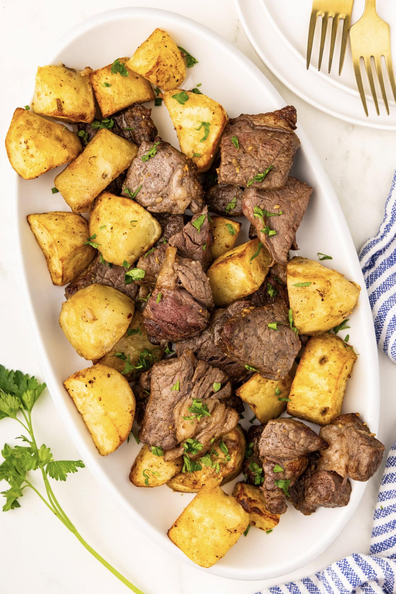 white platter with air fried potatoes and steak garnished with parsley, with plate and two forks in background