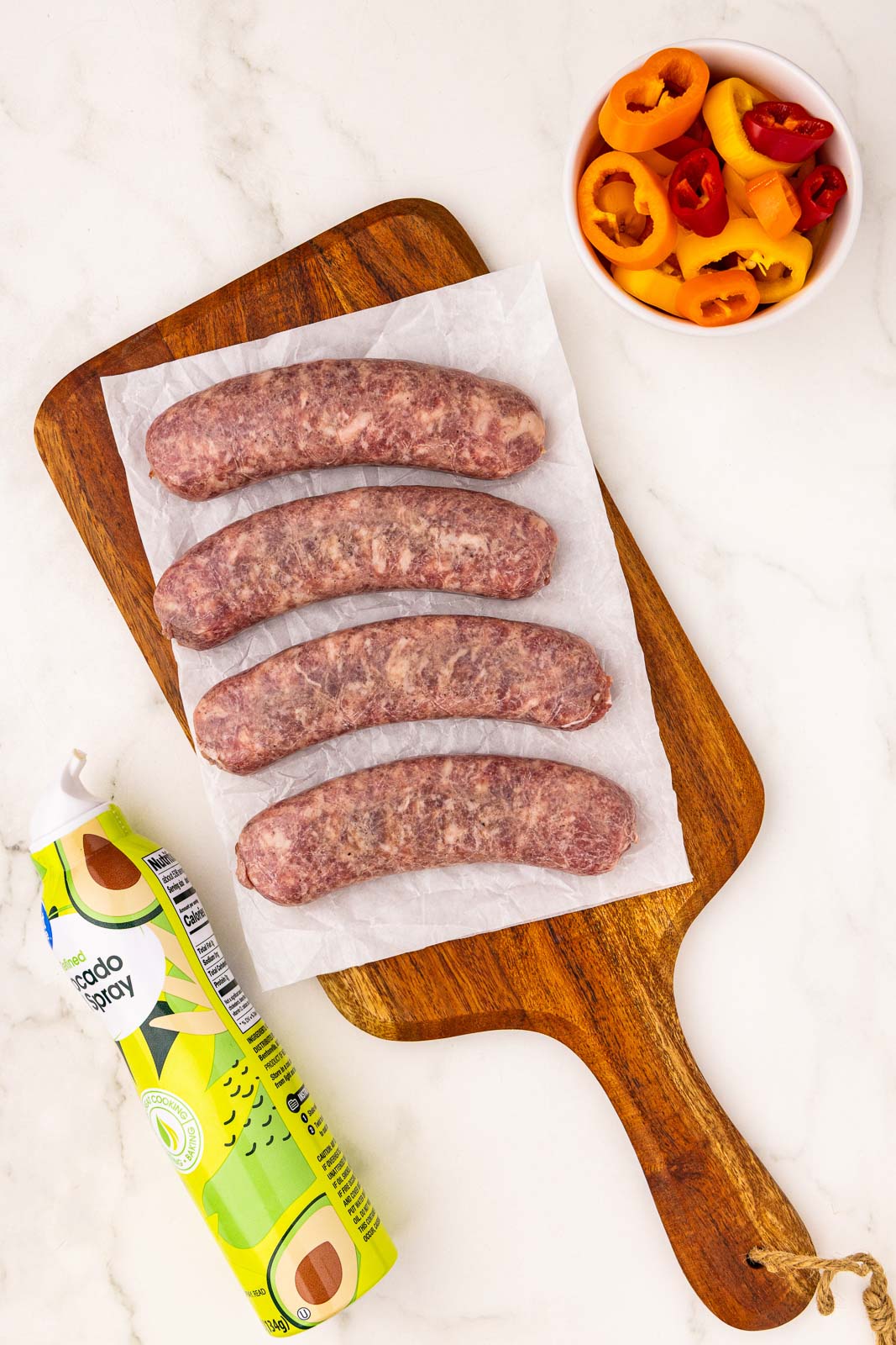 ingredients to include avocado spray, 4 uncooked brats on rectangular cutting board with white bowl of peppers