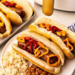 white plate with two air fried brats and peppers on buns, baked beans, Cole slaw, with slow cooker and tray of brats in background