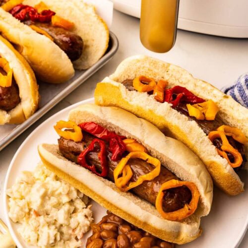 white plate with two air fried brats and peppers on buns, baked beans, Cole slaw, with slow cooker and tray of brats in background