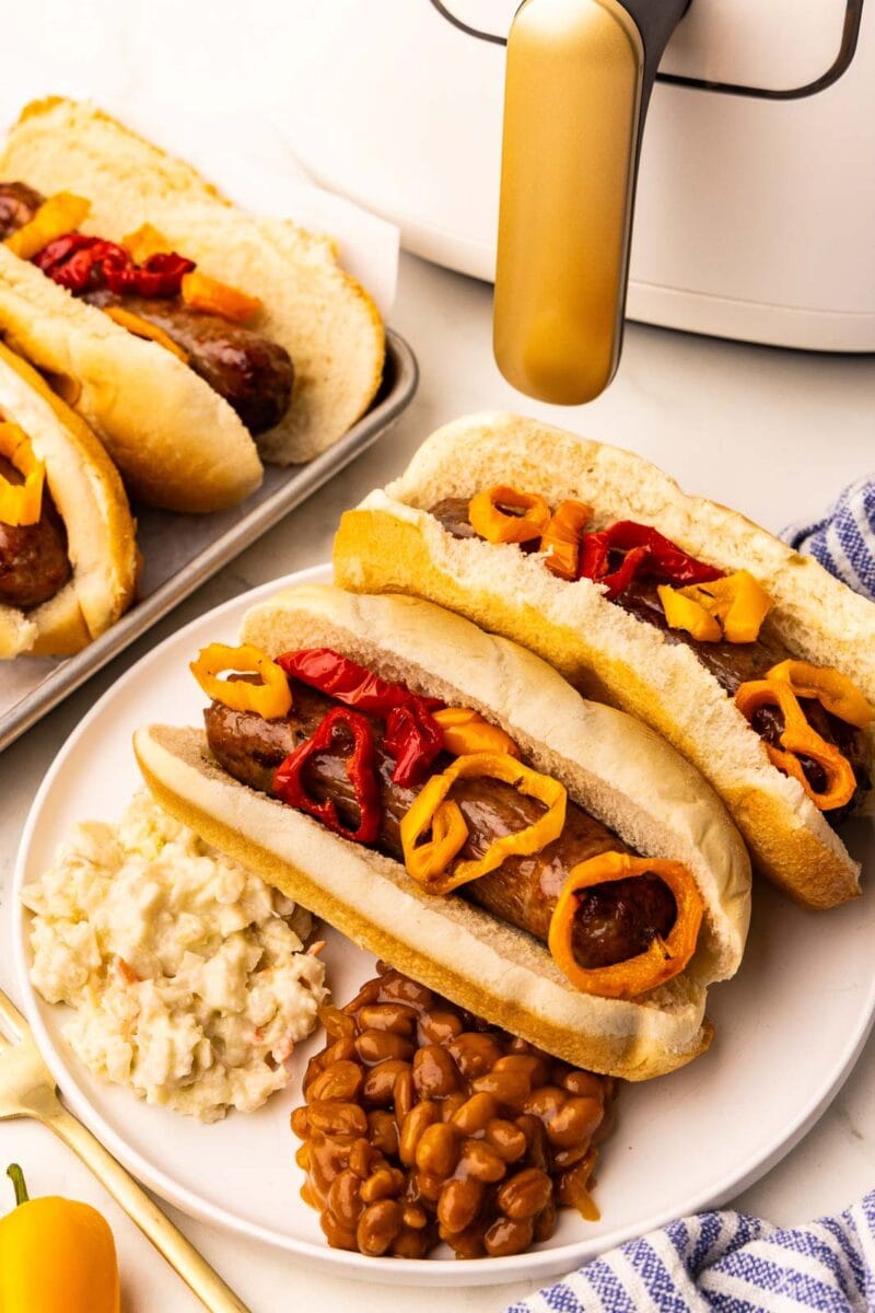 white plate with two air fried brats and peppers on buns, baked beans, Cole slaw, with slow cooker and tray of brats in background