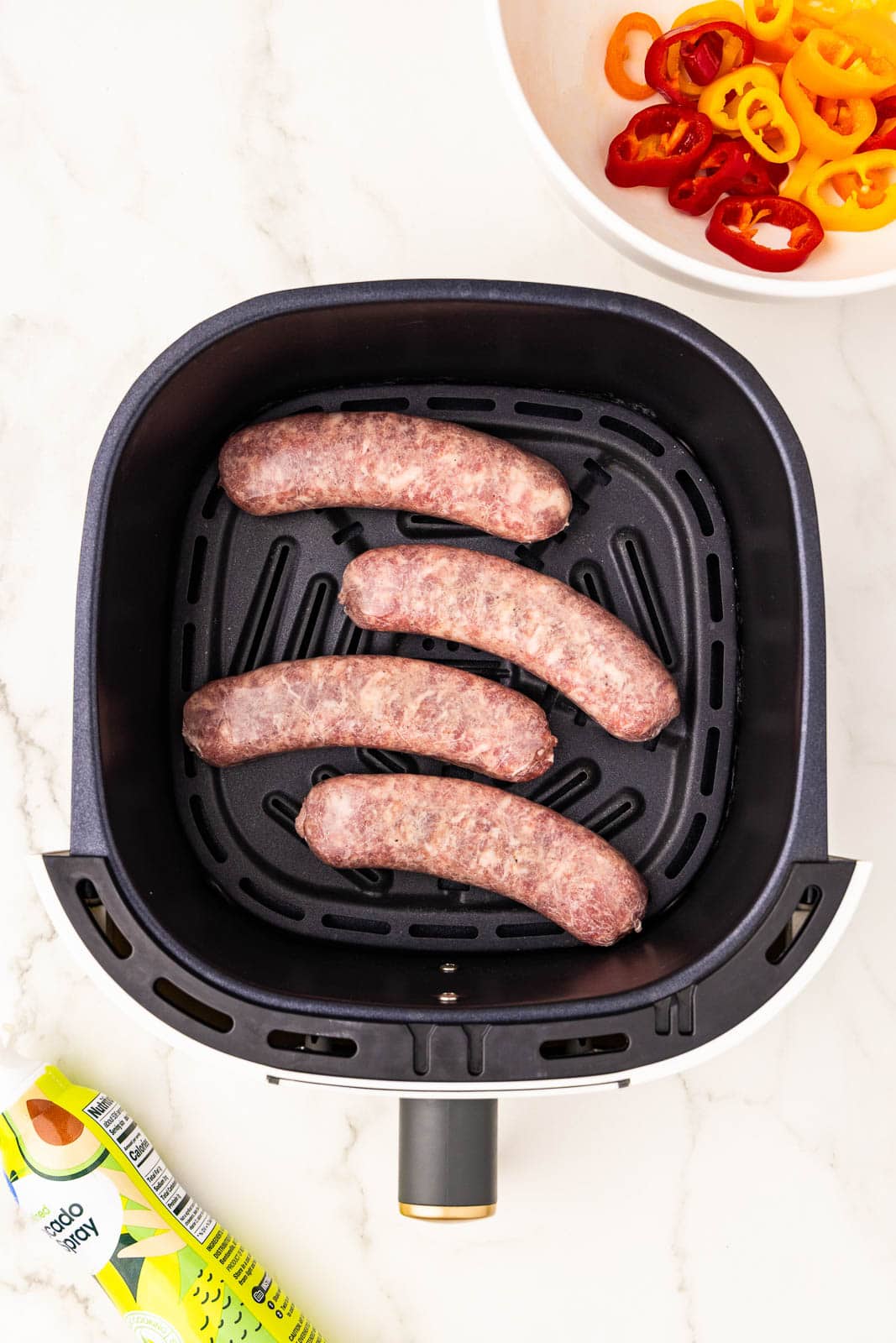 overhead shot of 4 uncooked brats in air fryer with white bowl of peppers in background