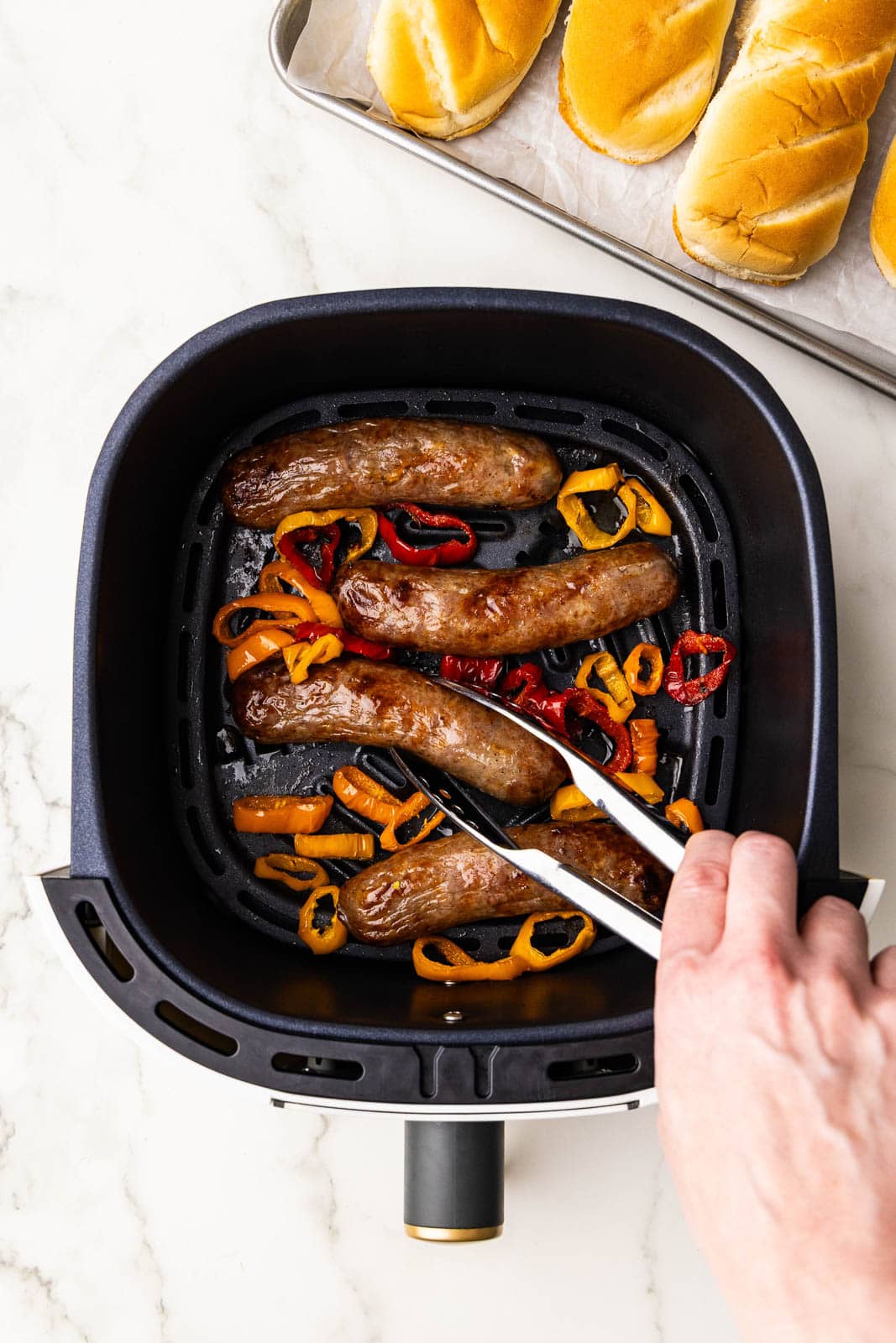 overhead shot of four brats in air fryer with peppers, with tray of buns in background