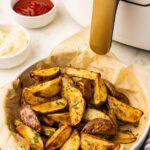 white bowl lined with parchment paper containing air fried potatoes, with ketchup, mayo and air fryer in background
