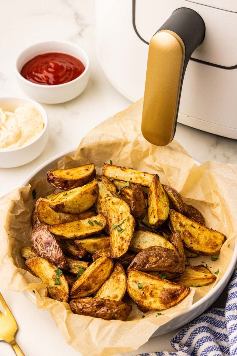 white bowl lined with parchment paper containing air fried potatoes, with ketchup, mayo and air fryer in background