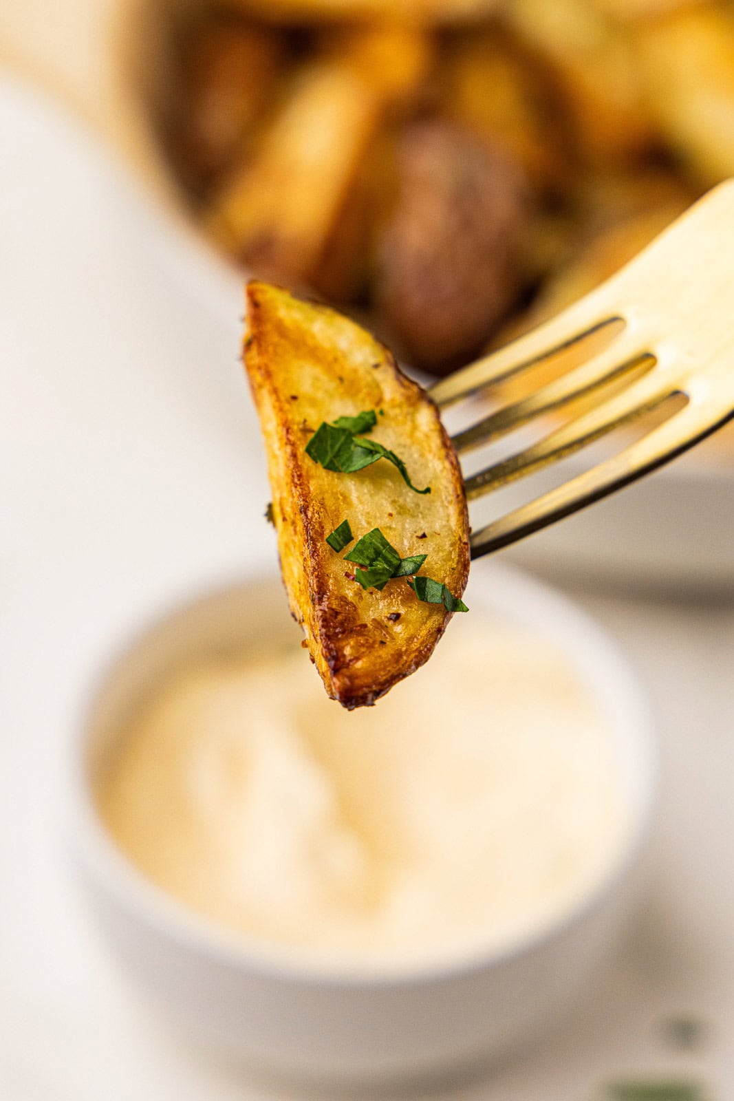 closeup of fork holding potato wedge with white bowl of mayo and bowl of potatoes in background