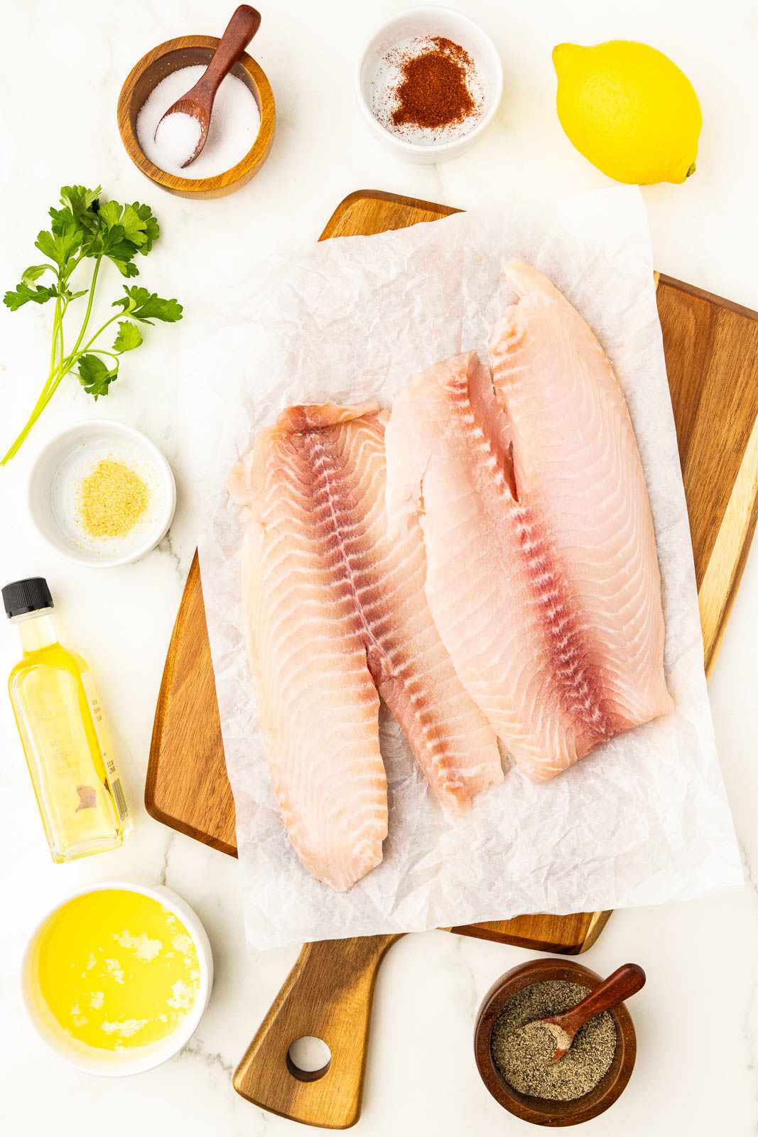 ingredients for air fryer tilapia to include 4 tilapia filet on wooden cutting board, surrounded by a bottle of olive oil, white bowl of melted butter, bowl of pepper, white bowl of garlic, bowl of paprika, bowl of salt, whole lemon, sprig of parsley