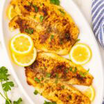 overhead shot of white platter with 4 tilapia filet, 3 slices of lemon and parsley on top, surrounded by blue and white napkin and sprig of parsley