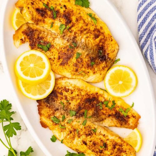 overhead shot of white platter with 4 tilapia filet, 3 slices of lemon and parsley on top, surrounded by blue and white napkin and sprig of parsley