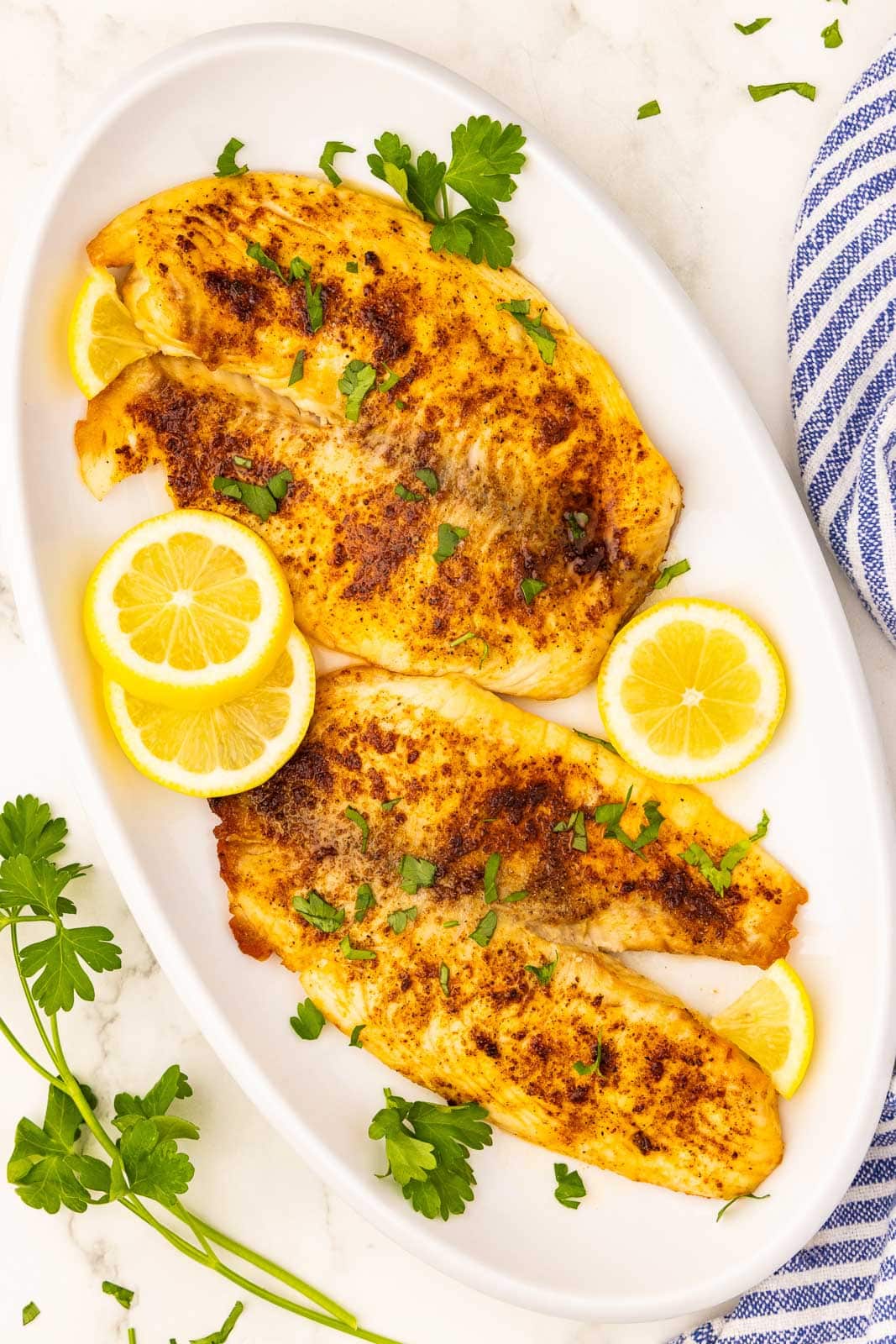 overhead shot of white platter with 4 tilapia filet, 3 slices of lemon and parsley on top, surrounded by blue and white napkin and sprig of parsley