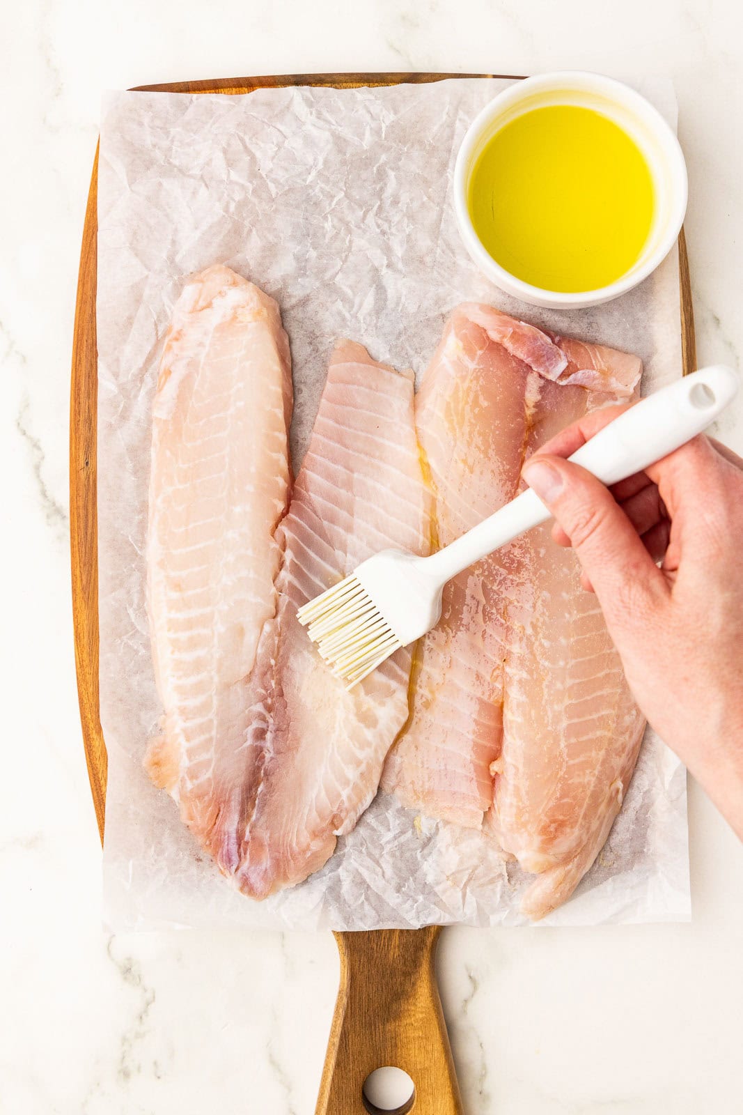 four tilapia filet on parchment lined wooden cutting board, small bowl of melted butter and tilapia brushed with butter with white brush