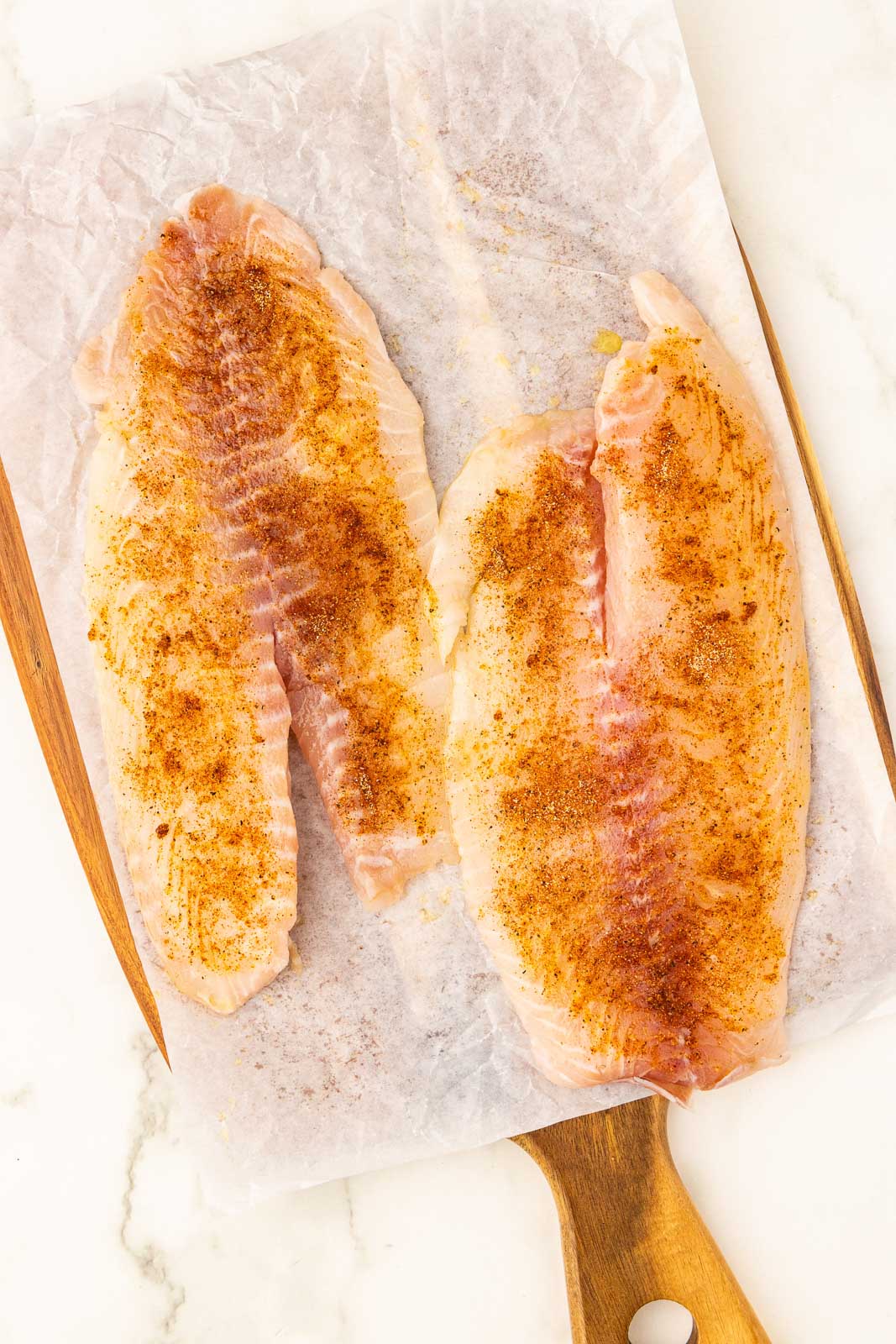4 tilapia filet on parchment lined wooden cutting board that have been seasoned with mixture
