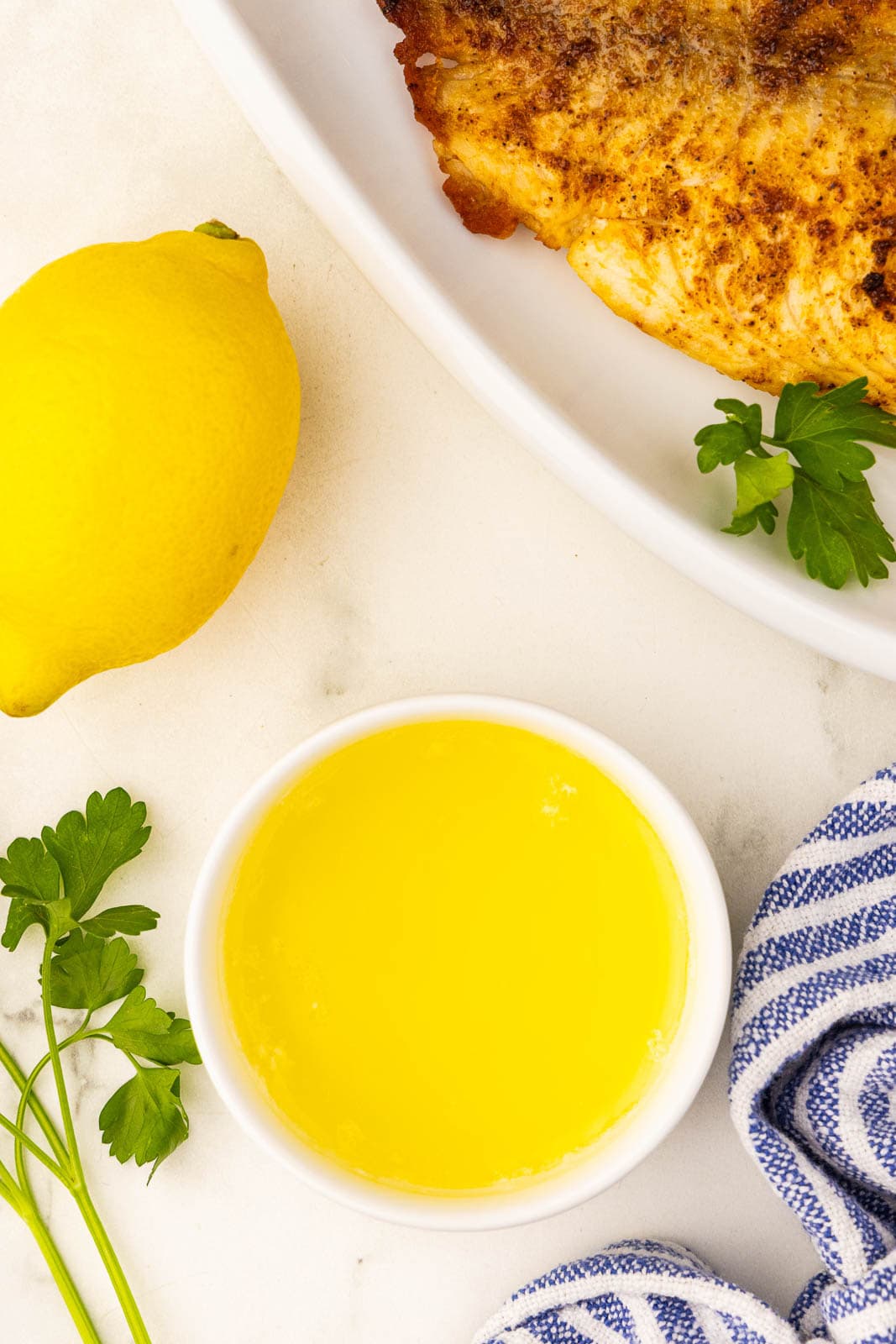 small bowl of melted butter, whole lemon, sprig of parsley, and small portion of dish of air fried tilapia