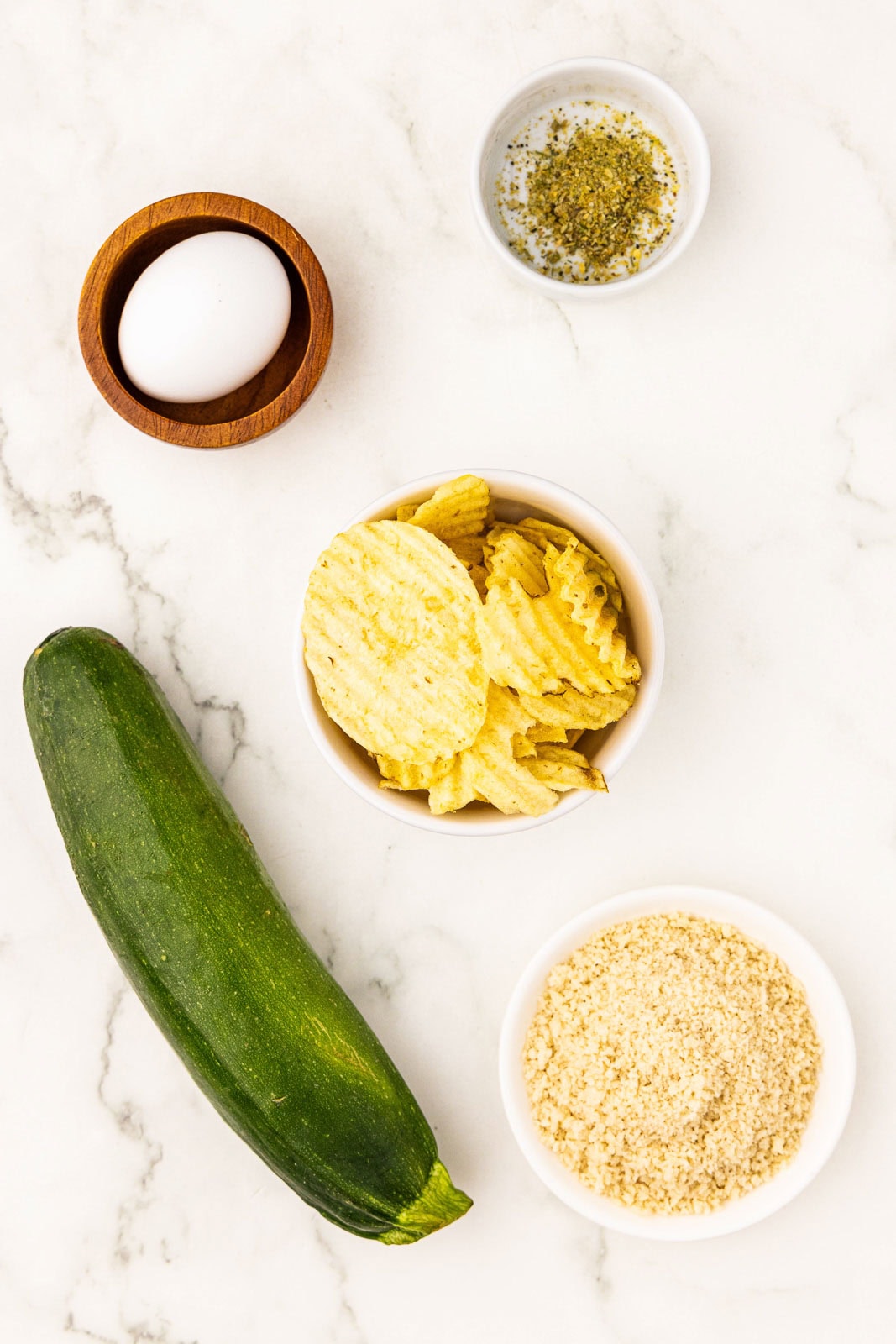 ingredients for air fryer zucchini fries to include one zucchini, small wooden bowl with egg, white bowl with seasoning, white bowl with potato chips, and white bowl with panko
