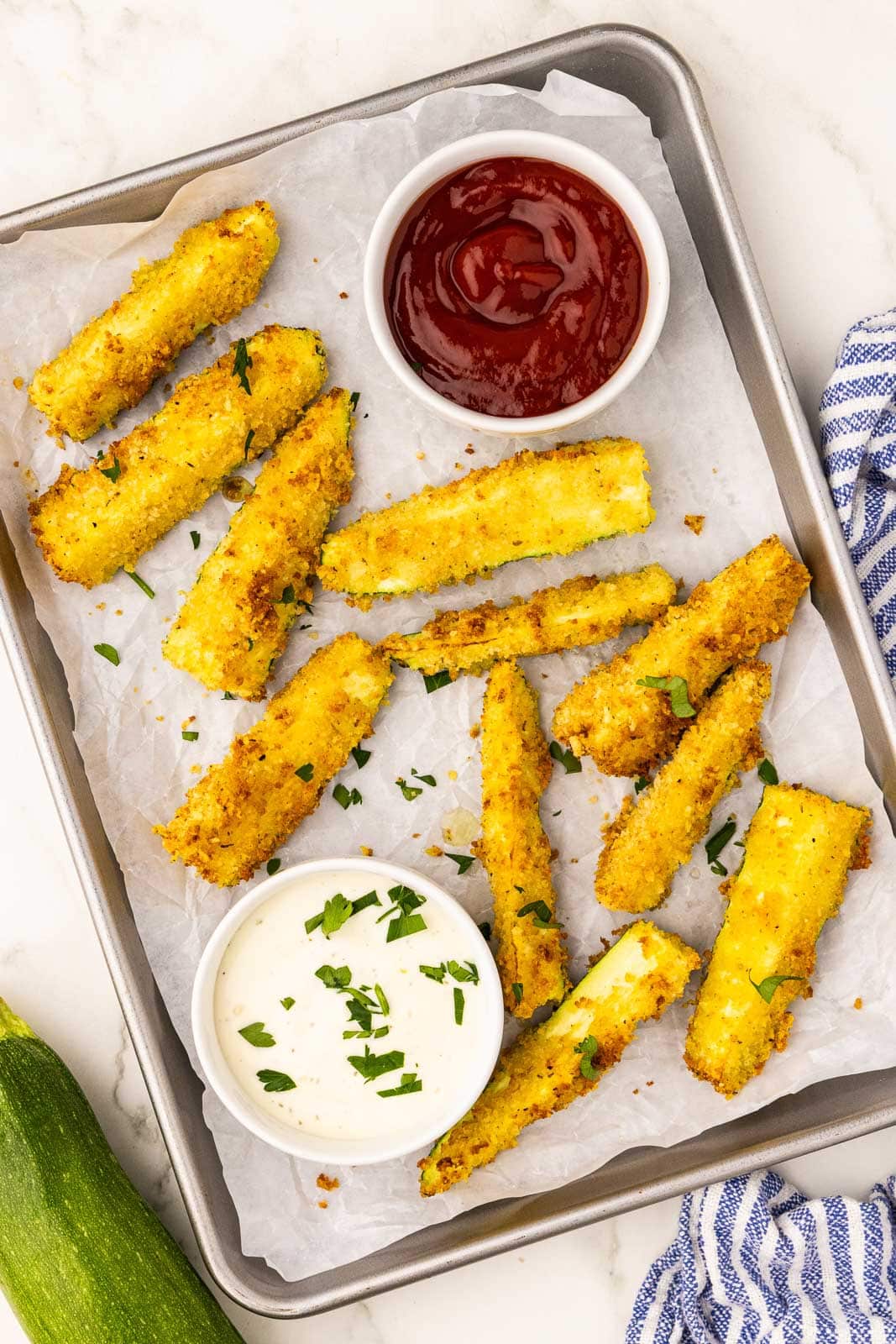 parchment lined cookie sheet containing air fried zucchini with two white bowls of ketchup and mayo