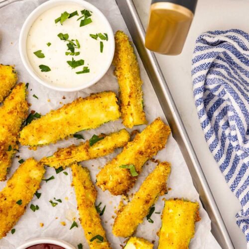 quarter view of parchment lined cookie sheet containing air fried zucchini with two white bowls of ketchup and mayo with air fryer and blue and white dish towel in background