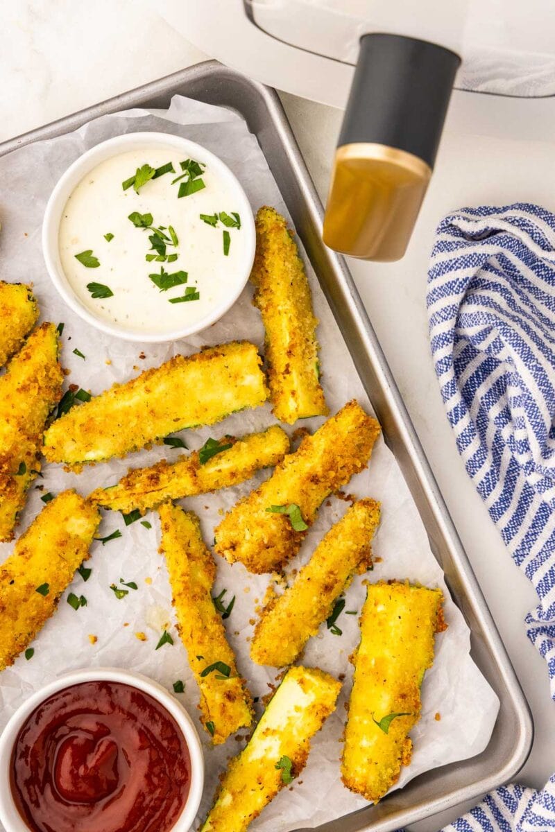 quarter view of parchment lined cookie sheet containing air fried zucchini with two white bowls of ketchup and mayo with air fryer and blue and white dish towel in background
