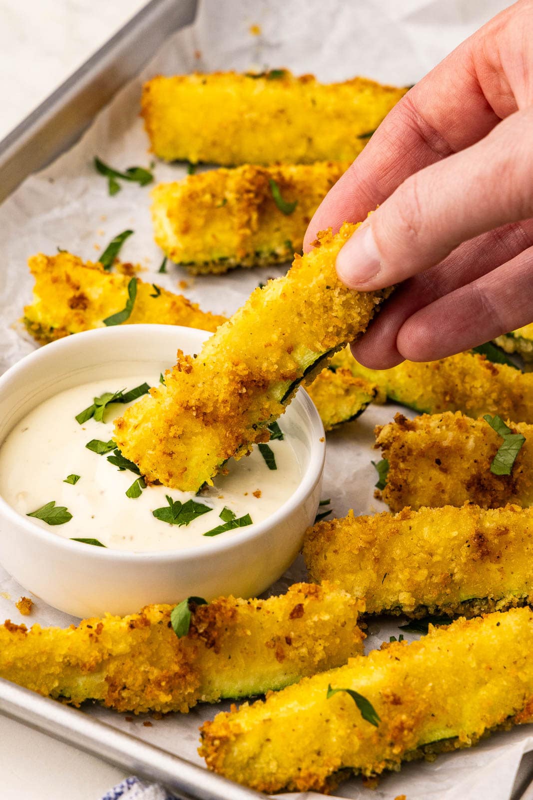 closeup view of cookie sheet lined by parchment paper with zucchini fries with a hand dipping into a small bowl of mayo