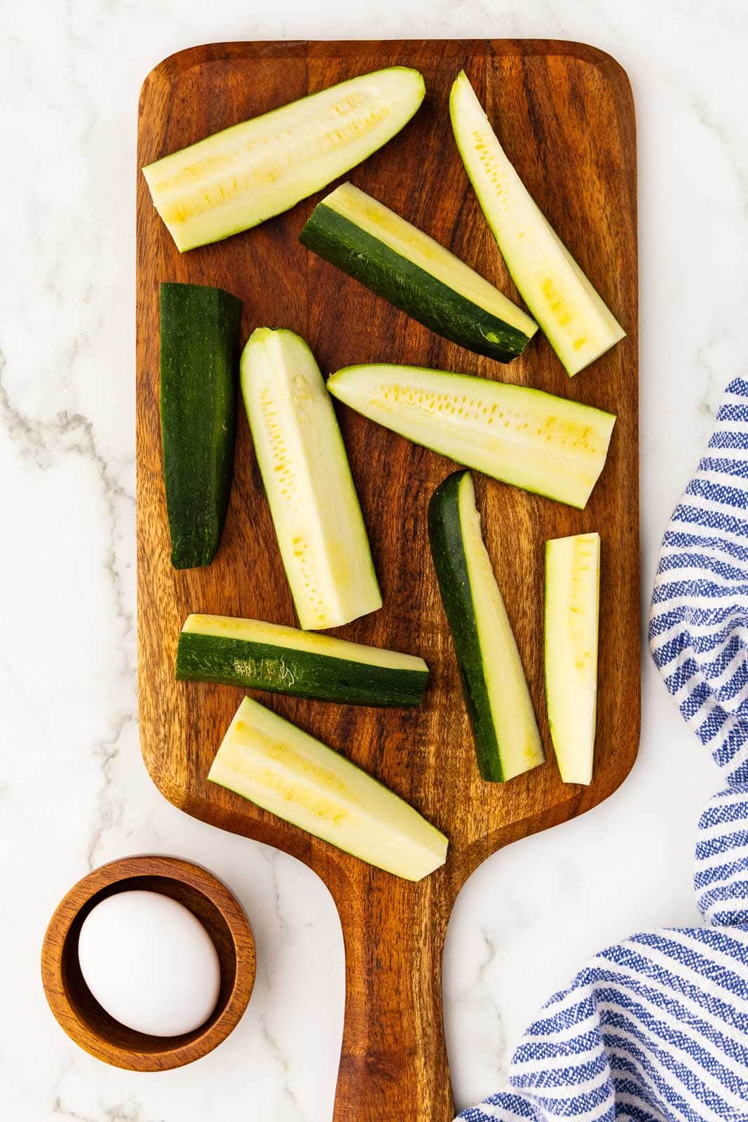 wooden cutting board with sliced zucchini, surrounded by wooden bowl with egg and blue and white striped dish towel
