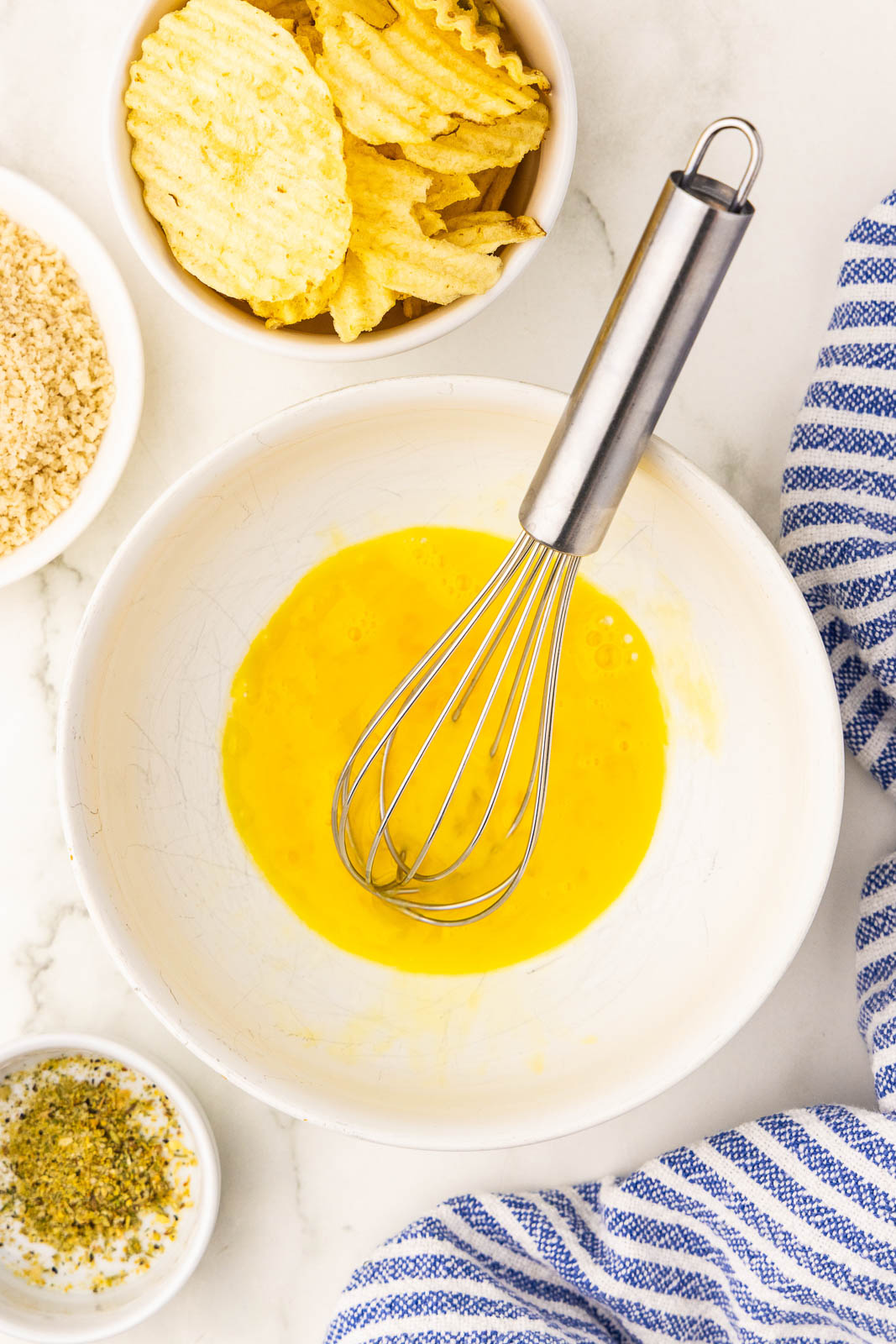 white bowl with beaten egg with silver whisk, surrounded by small white bowls of potato chips, panko, seasoning and blue and white striped dish towel
