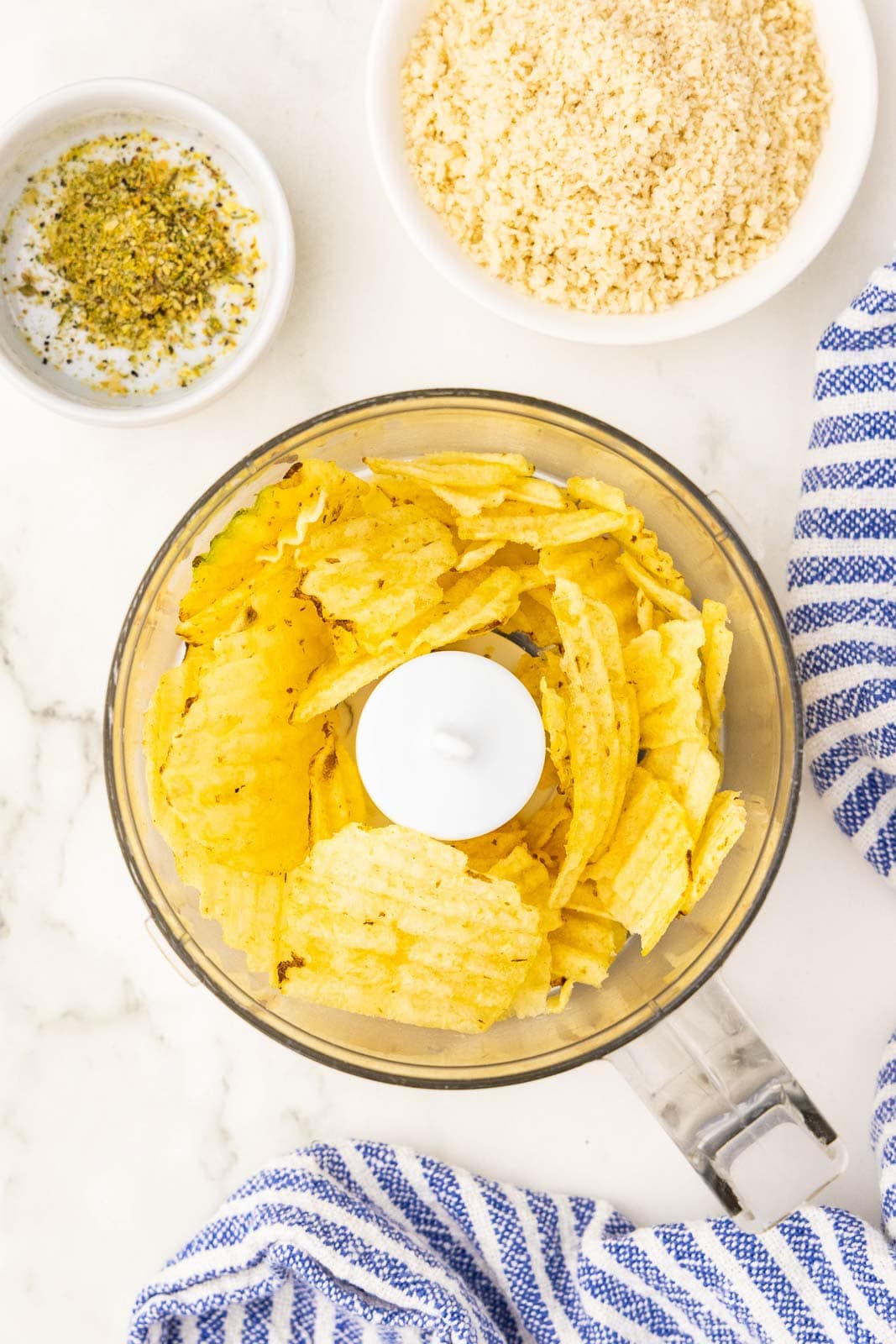 overhead view of small food processor holding potato chips, with small bowls of seasoning and panko with blue and white dish towel on side