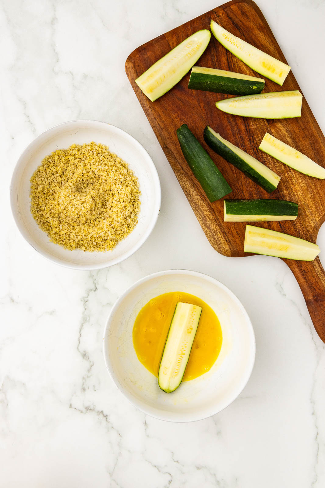 wooden cutting board with sliced zucchini, with white bowls containing panko mixture and beaten egg with zucchini